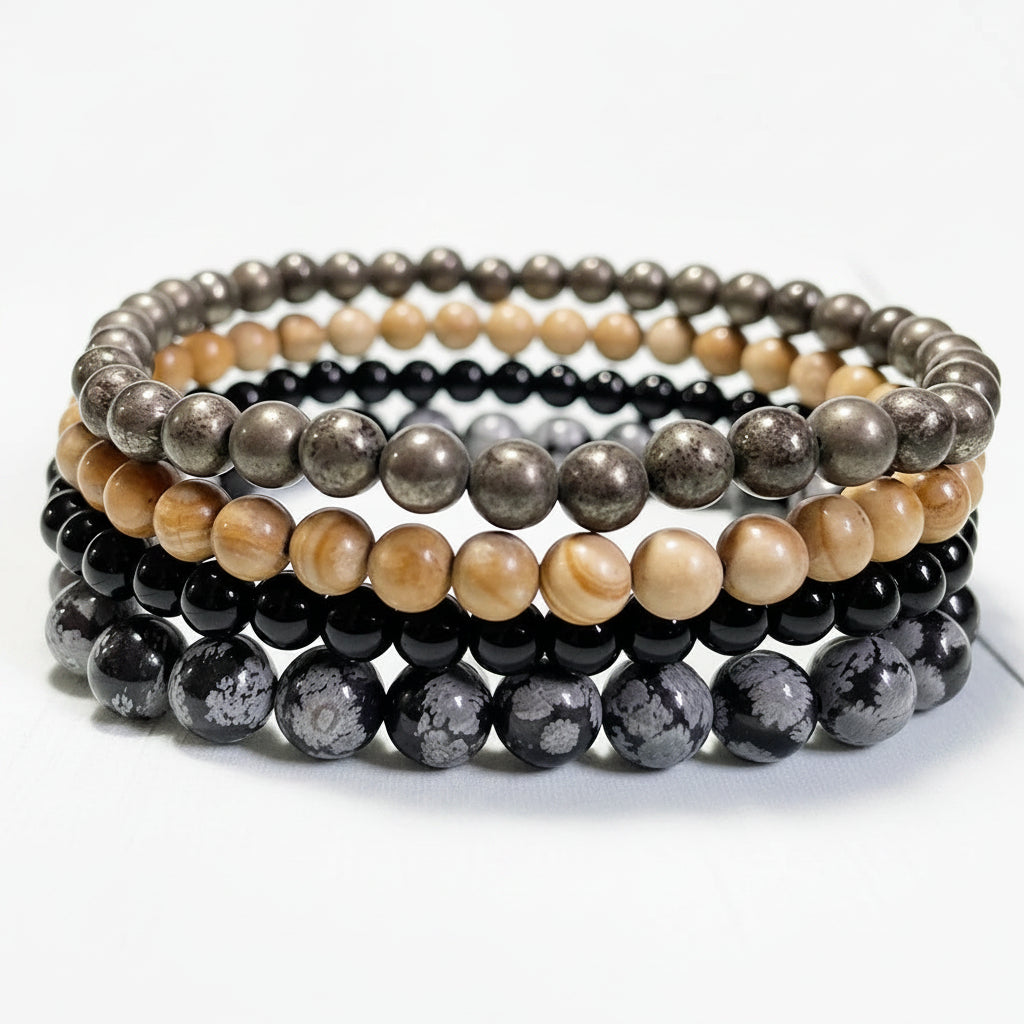 Stack of beaded bracelets of pyrite, tourmaline, snowflake obsidian, and jasper on a white surface.