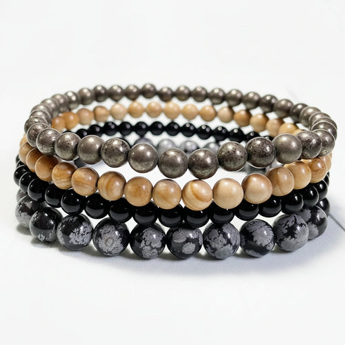 Stack of beaded bracelets of pyrite, tourmaline, snowflake obsidian, and jasper on a white surface.