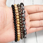 Set of four beaded bracelets of tourmaline, jasper, pyrite, and osidian on a hand against a light background.